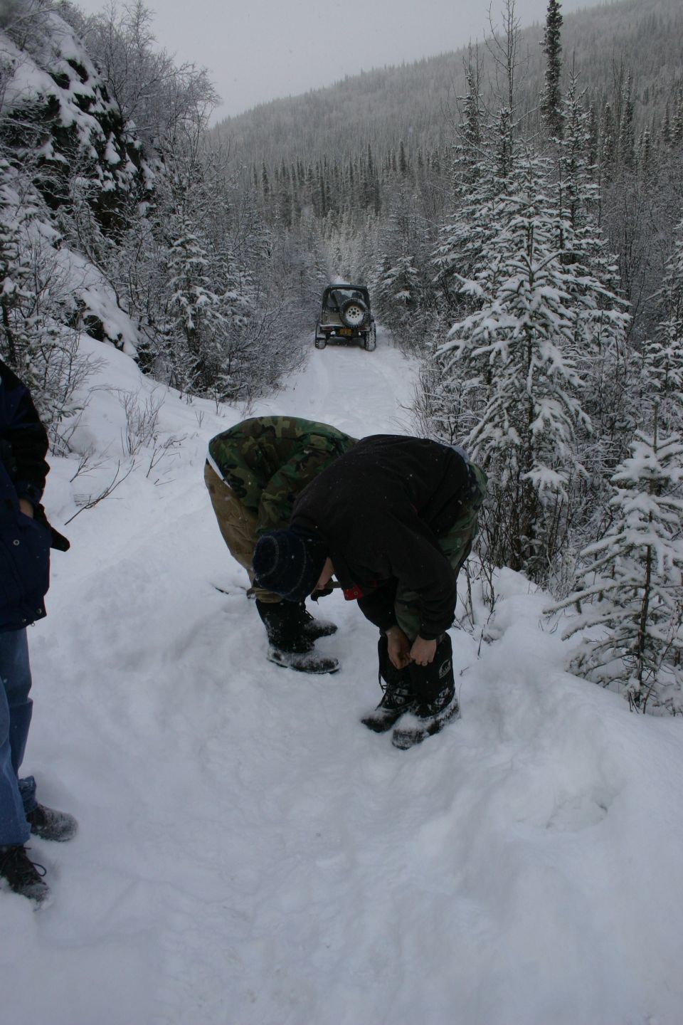 Two people adjusting boots on a snowy trail with a Jeep Wrangler in the distance