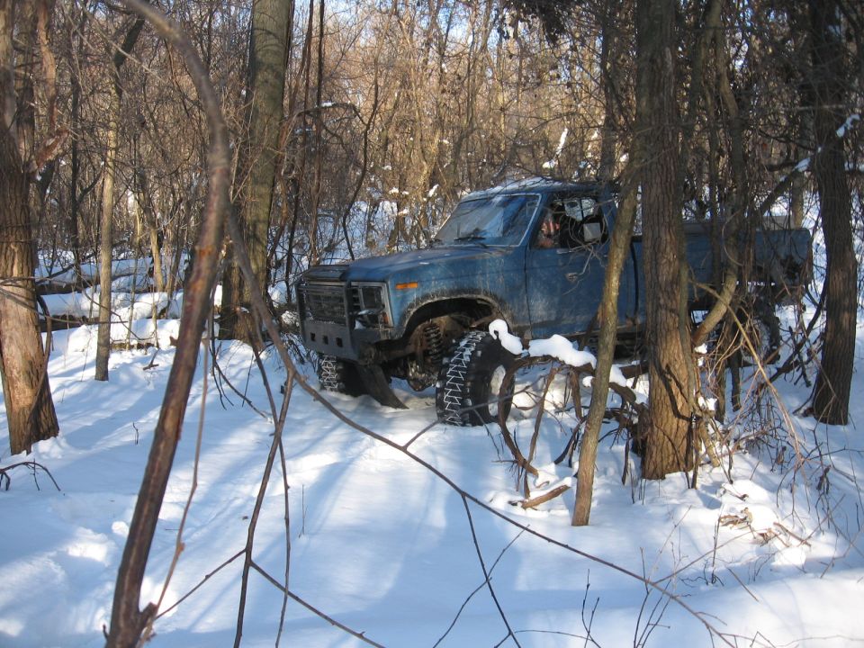 Blue pickup truck with off-road tires in snow-covered woods. Aftermarket bumper visible