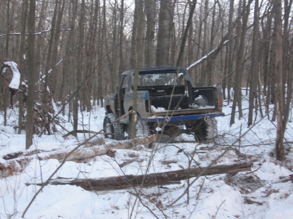 Blue pickup truck with large tires stuck in snow, surrounded by trees and fallen logs