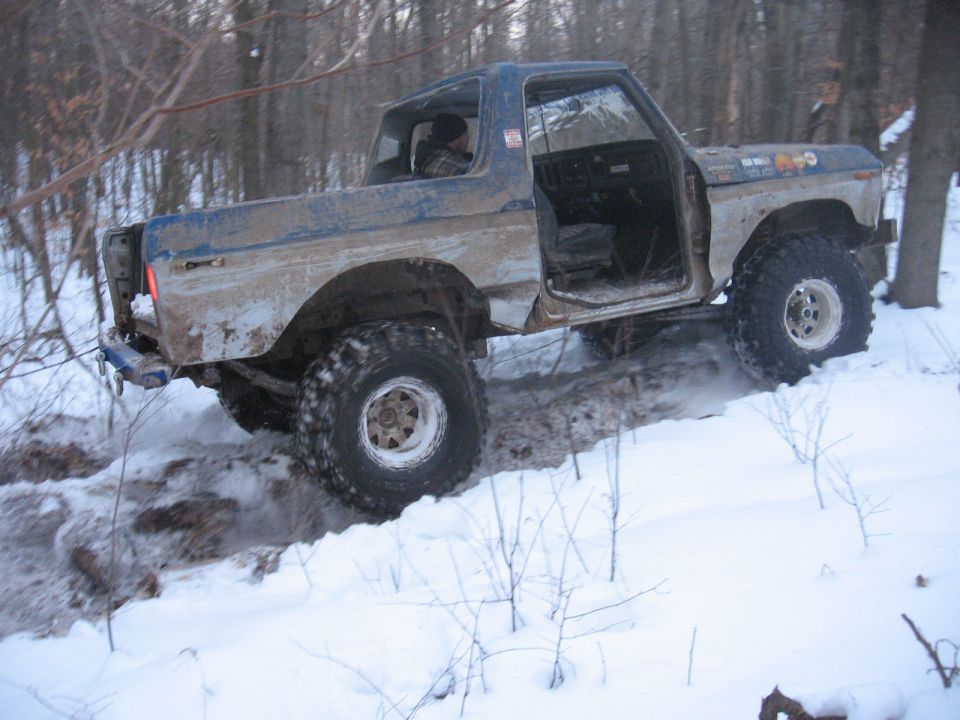 Blue Ford Bronco with doors open driving through snow and mud in a wooded area. Some stickers on the hood