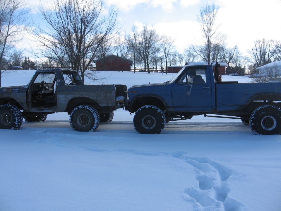Gray Ford Bronco and blue Ford pickup truck parked side-by-side in snow, both lifted with large tires