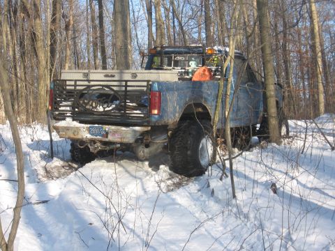 1979 Ford Bronco - A lifted blue pickup truck navigates through a snowy wooded area, its large tires digging into the snow.