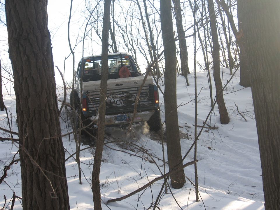 White pickup truck with a black stripe on the tailgate, driving through snow in a wooded area