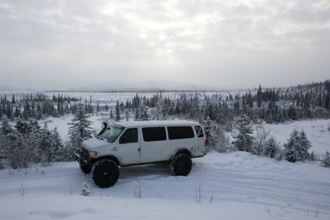 2003 Jeep TJ - A white van equipped with large off-road tires sits in a snow-covered landscape with pine trees and a cloudy sky.