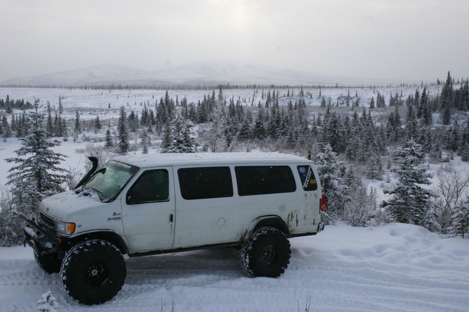 White van with big off-road tires parked in a snowy landscape with pine trees and mountains in the background