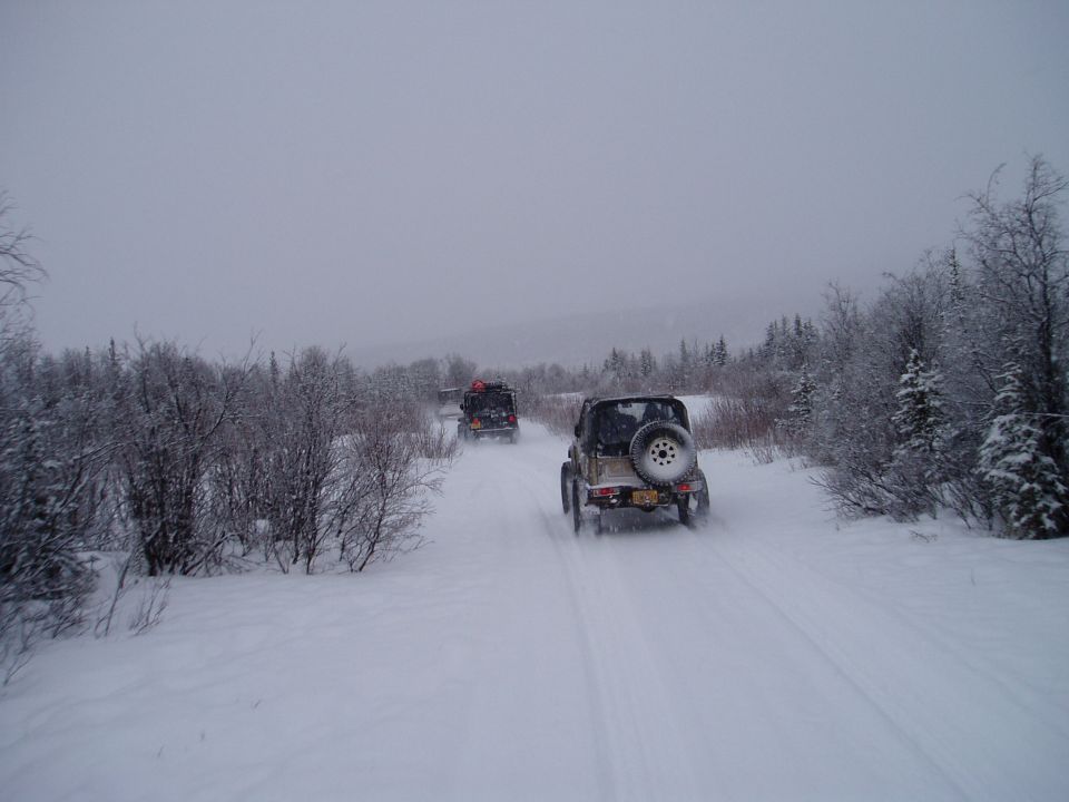 Tan Jeep Wrangler with a spare tire on the back drives on a snowy road behind another vehicle