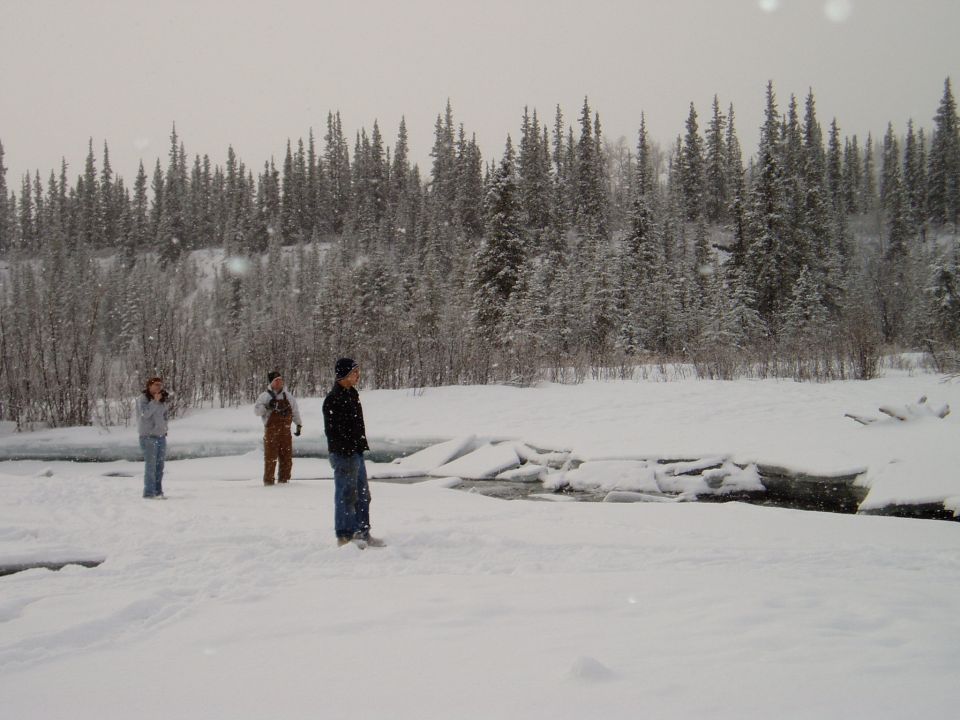 Three people stand in a snowy field near a partially frozen river with a dense forest in the background