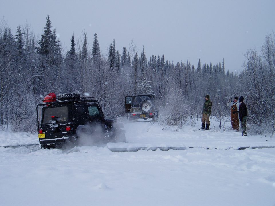 Black Jeep Wrangler with roof rack and red cargo box driving through snow, rear view