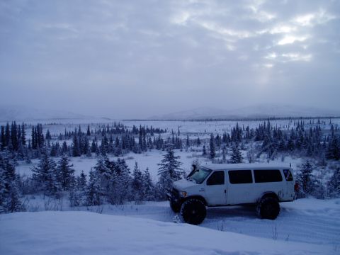 2000 Ford E350 - A white 2000 Ford E350 van equipped with large off-road tires sits in a snowy, wooded landscape under a cloudy sky.