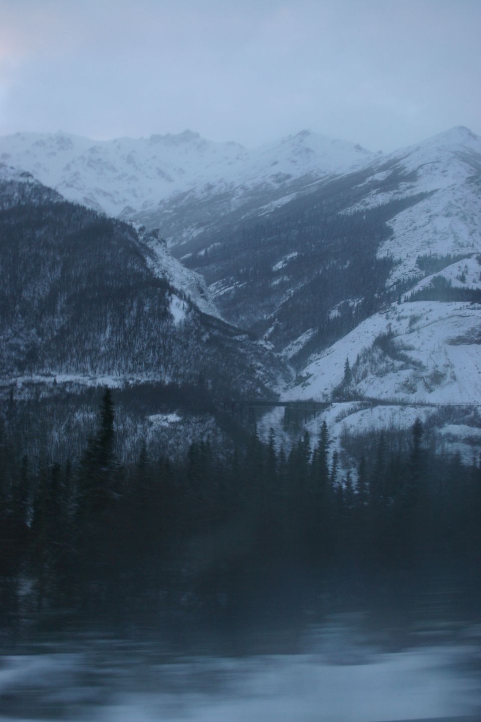 Snowy mountains reflected in a dark, still lake with pine trees in the foreground
