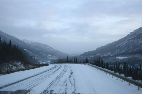 2003 Jeep TJ - A winding road covered in snow leads through a valley with snow-covered mountains and pine trees under a cloudy sky.