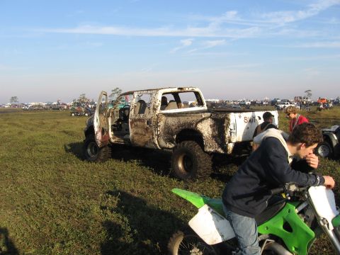 1974 Ford F-100 - A white crew-cab pickup stripped of glass and doors, caked in dried mud and lifted on oversized mud tires, sits in a grassy field at a mud-…