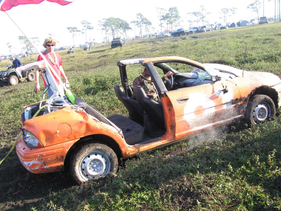 Orange stripped-out beater sedan with roof and doors cut off, hood open, stuck in a grass field
