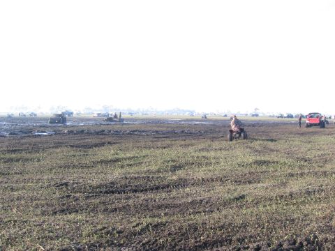 1974 Ford F-100 - Hazy backlit view across a wide, rutted mud-bog field with deep tire tracks cutting through patchy grass. A rider on a red ATV crosses the…