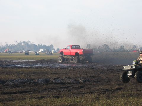 1974 Ford F-100 - A red lifted GM-style regular-cab pickup on tall mud tires blasts through a churned mud pit, throwing a huge rooster tail of black mud into…