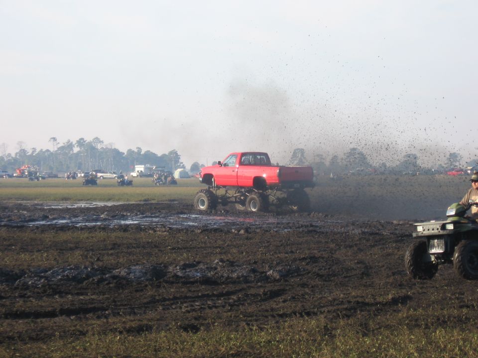Red lifted pickup slinging a rooster tail of mud through a mud pit at an off-road event