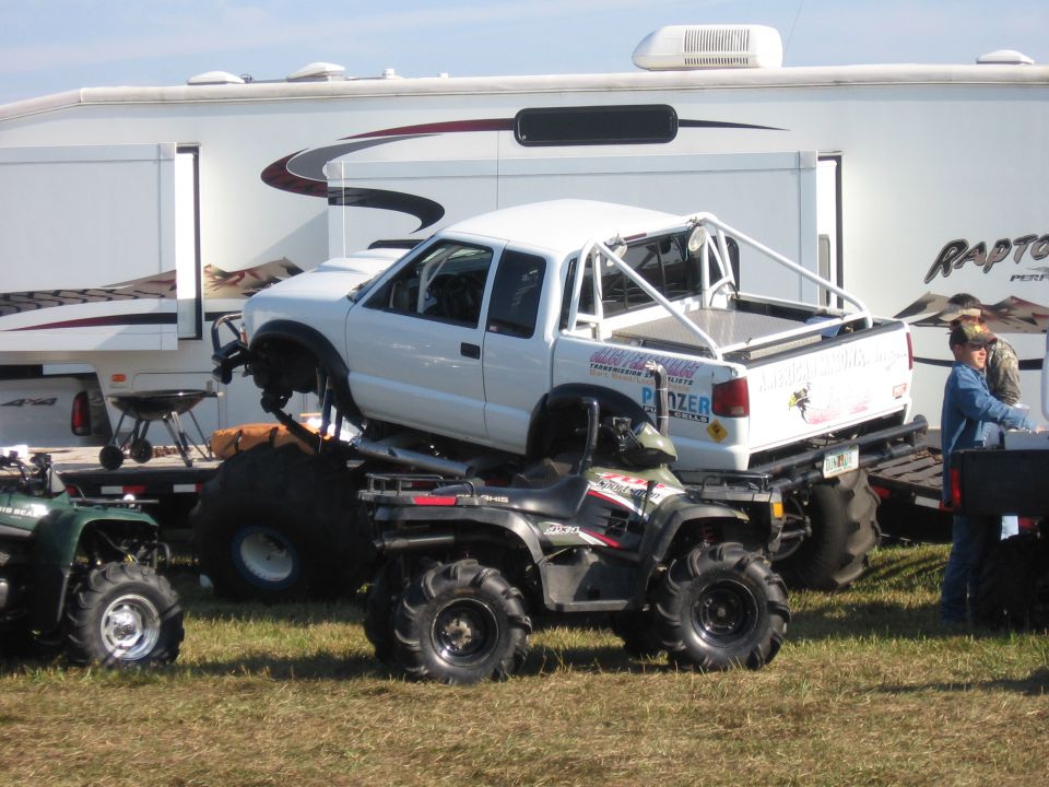 White caged mud-race pickup with big tires on a trailer beside a Raptor toy hauler and ATVs