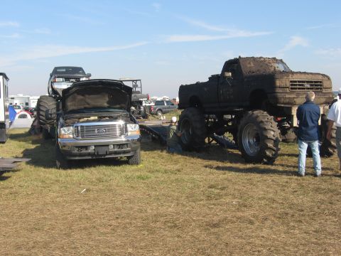 1974 Ford F-100 - Pit scene at a grassy mud event: a dark Ford Super Duty tow truck sits hood-up beside a gooseneck trailer, while a mud-caked lifted 1990s F…