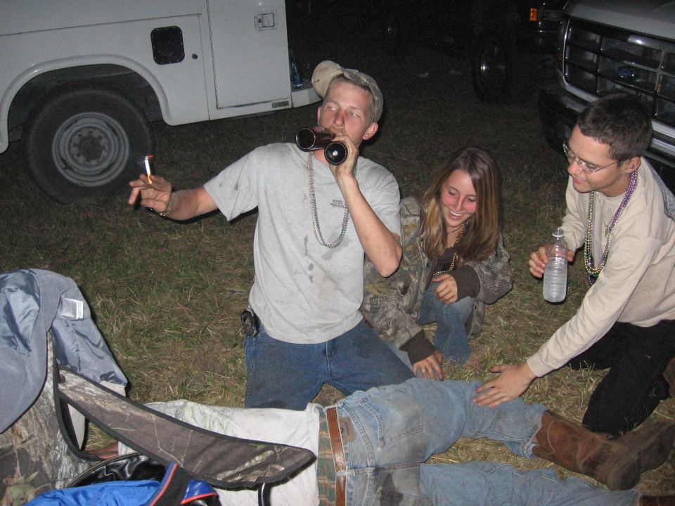 Two trucks, one white utility truck and a silver pickup truck, are parked on grass at night with people gathered around