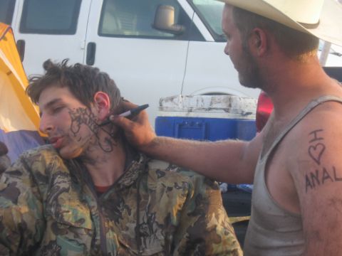 1974 Ford F-100 - Campsite horseplay at an outdoor event: one man in a straw cowboy hat and tank top draws on the face of another man asleep in a camouflage…