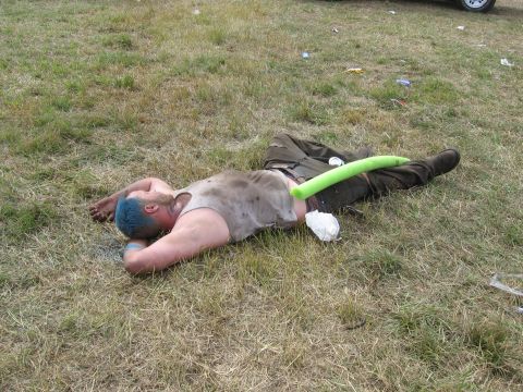 1974 Ford F-100 - A person with blue-dyed hair, a dirty tank top and cargo pants lies passed out on trampled grass with a green foam pool noodle across their…