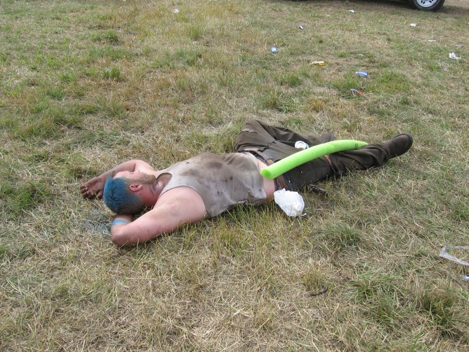 Person lying asleep on trampled grass with a green pool noodle at an outdoor event