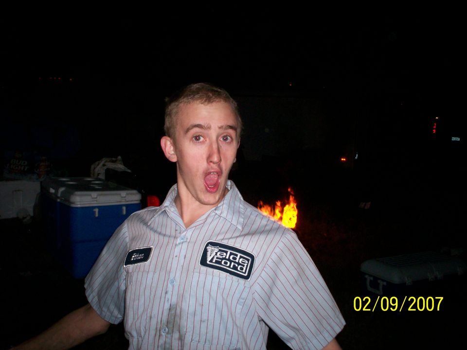 Man wearing a striped shirt with a Velde Ford patch, standing in front of a fire at night