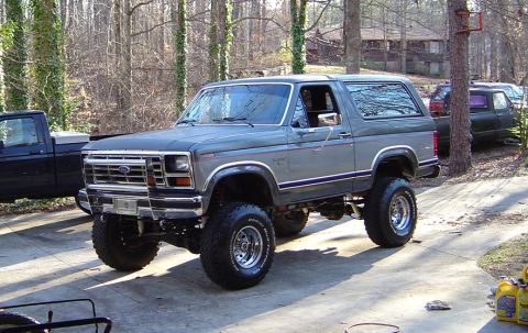 1986 Ford Bronco - A two-tone gray 1980s full-size Ford Bronco with removable hardtop sits three-quarter front on a concrete driveway, lifted on chrome modula…