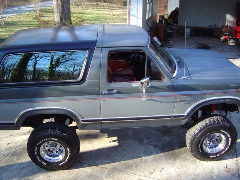 1986 Ford Bronco - Driver-side profile of a lifted two-tone gray Ford Bronco with a removable hardtop, red pinstripe and red interior, parked on a concrete dr…