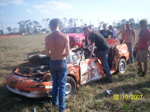 1974 Ford F-100 - A group of people examine a severely damaged orange car in a grassy field.