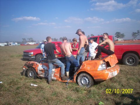 1974 Ford F-100 - Several men gather around a heavily damaged orange car, with red trucks in the background. The photo is dated 02/10/2007.
