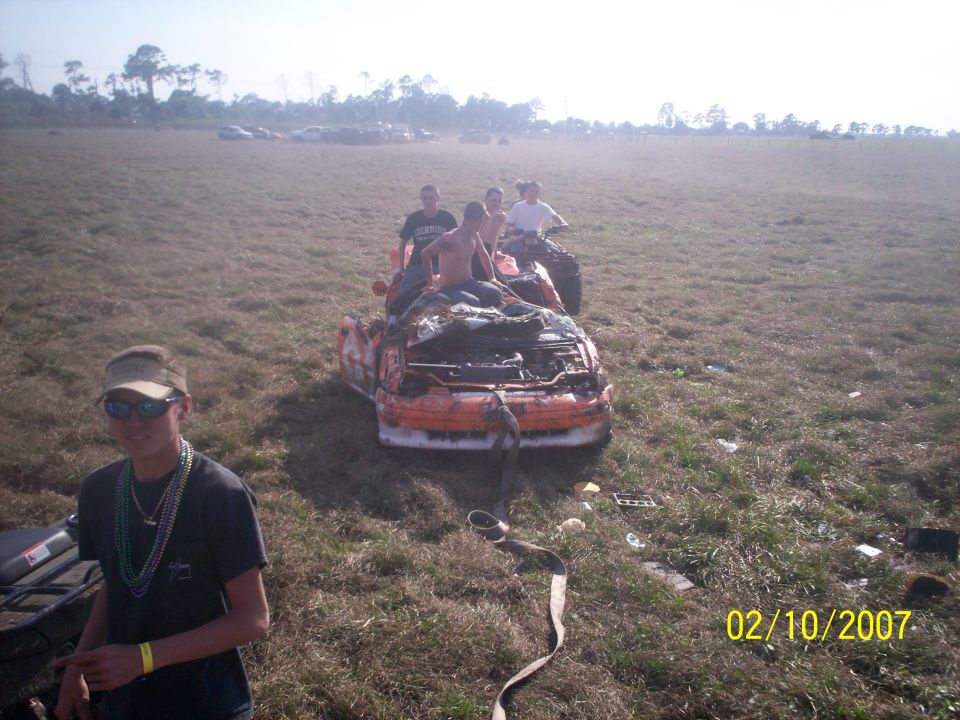 Wrecked orange car with people riding on top, being towed in a field