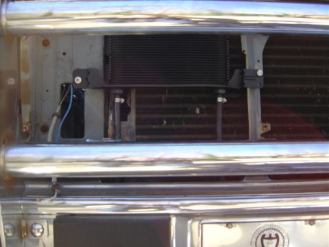 1986 Ford Bronco - Close-up through chrome front-end bars showing a black plate-style auxiliary cooler mounted on brackets ahead of the condenser fins, plumbe…