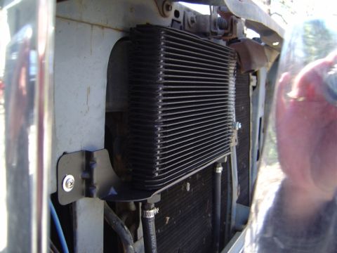 1986 Ford Bronco - Close-up of a black finned transmission or oil cooler mounted with a black metal bracket held by a Phillips-head bolt in front of a radiato…
