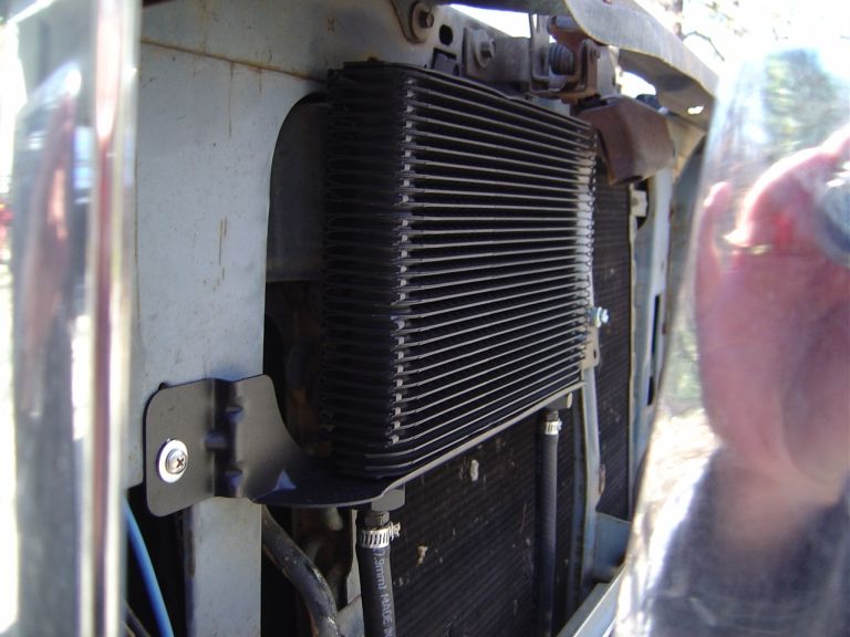 Black finned oil cooler mounted with bracket in front of a vehicle radiator