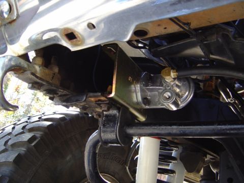 1986 Ford Bronco - Underside view behind the front bumper showing a gold zinc-plated bracket bolted to the frame rail holding a remote spin-on filter mount wi…