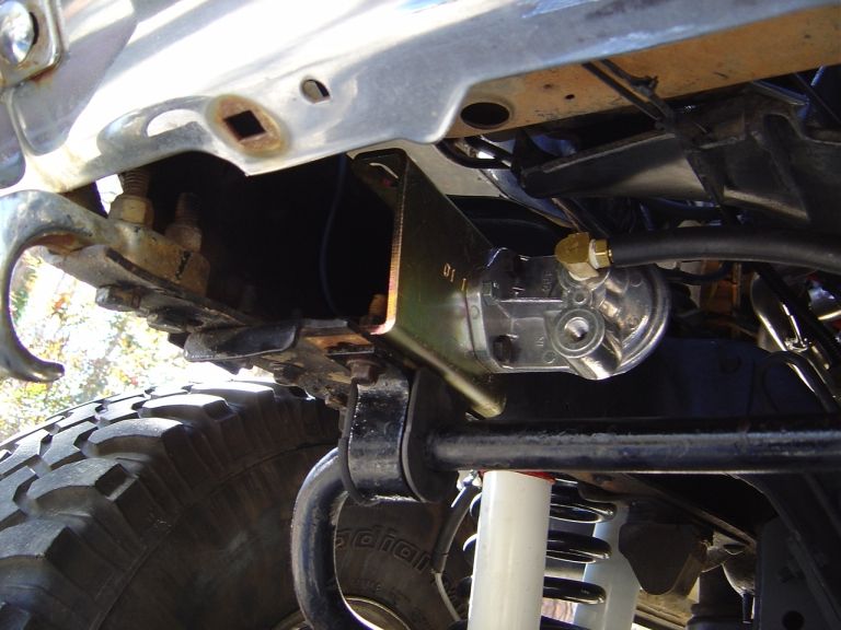 Frame-mounted remote oil filter head on gold bracket above white shock and mud tire