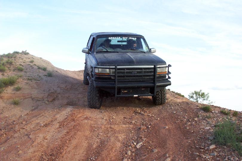 Dark blue Ford Bronco with brush guard climbing a red dirt hill against the sky