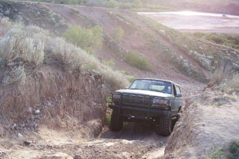 1995 Ford Bronco - A dark green Ford Bronco navigates a narrow dirt path with dry brush and hills in the background.