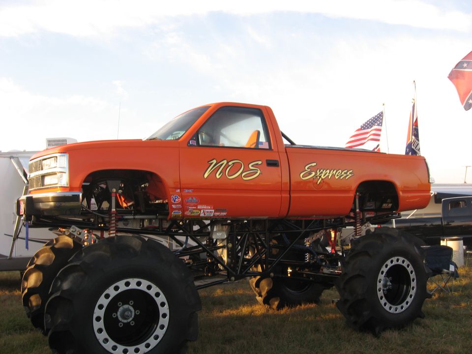 Orange 'NOS Express' tube-chassis GM mud truck on tractor tires parked on grass by trailers