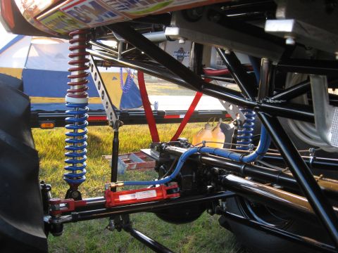 1974 Ford F-100 - Close-up under a tube-chassis mud/race truck showing the front suspension: a coilover with stacked red and blue dual-rate springs on an adj…