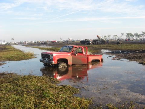 1974 Ford F-100 - A red square-body Chevrolet/GMC regular-cab pickup with a windshield banner sits axle-deep in standing water and mud at an off-road park, i…
