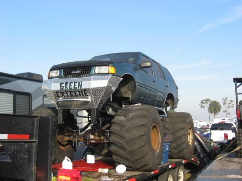 1974 Ford F-100 - Teal Isuzu Rodeo built as a mini monster truck, sitting on a black flatbed trailer with its front three-quarter side facing the camera. It…