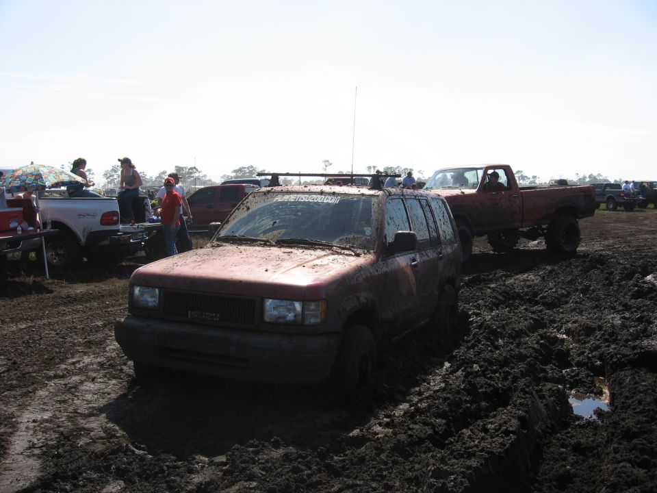 Muddy red SUV stuck in mud pit with red pickup truck nearby. People watching