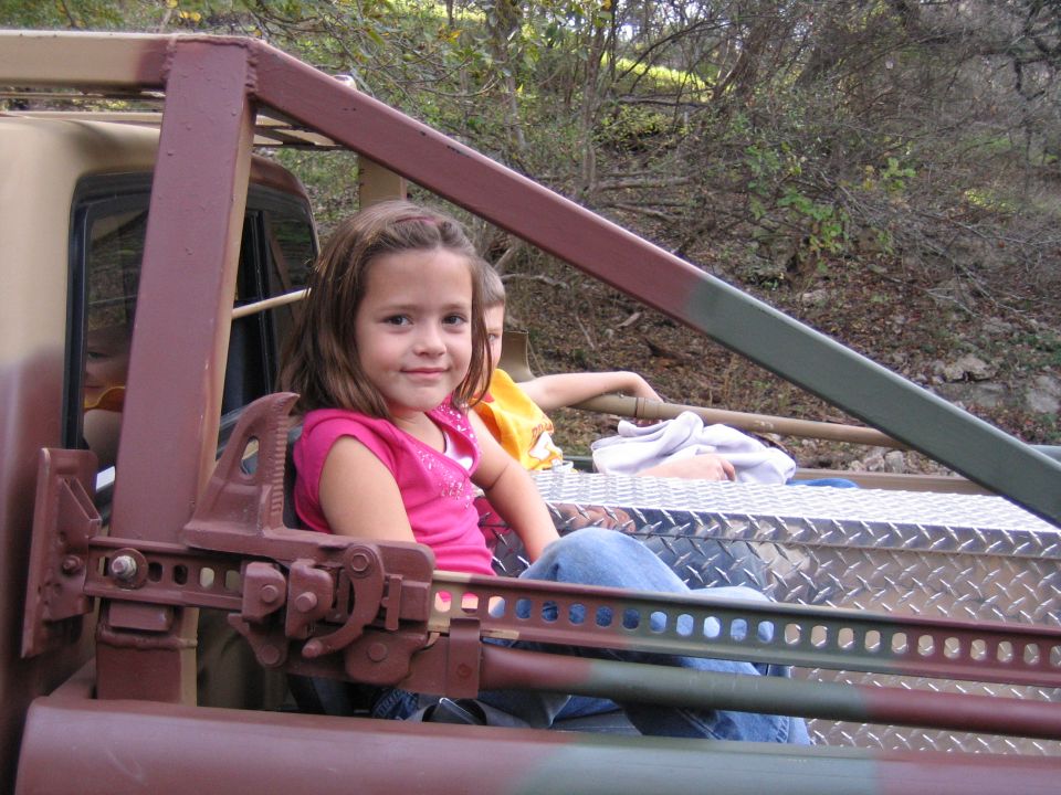 Two kids sit in the bed of a camouflaged truck with a roll cage