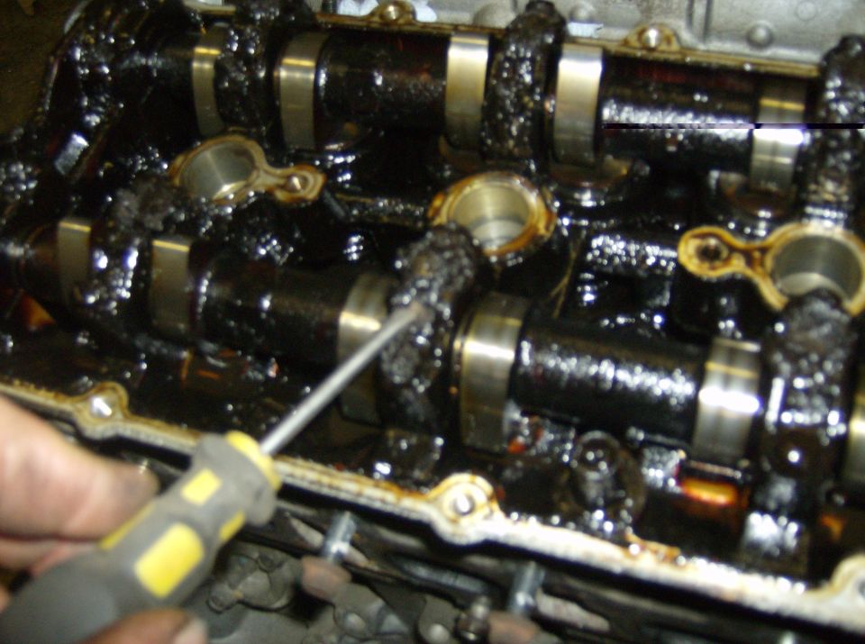Sludged DOHC cylinder head with camshafts exposed, screwdriver pointing at black deposits