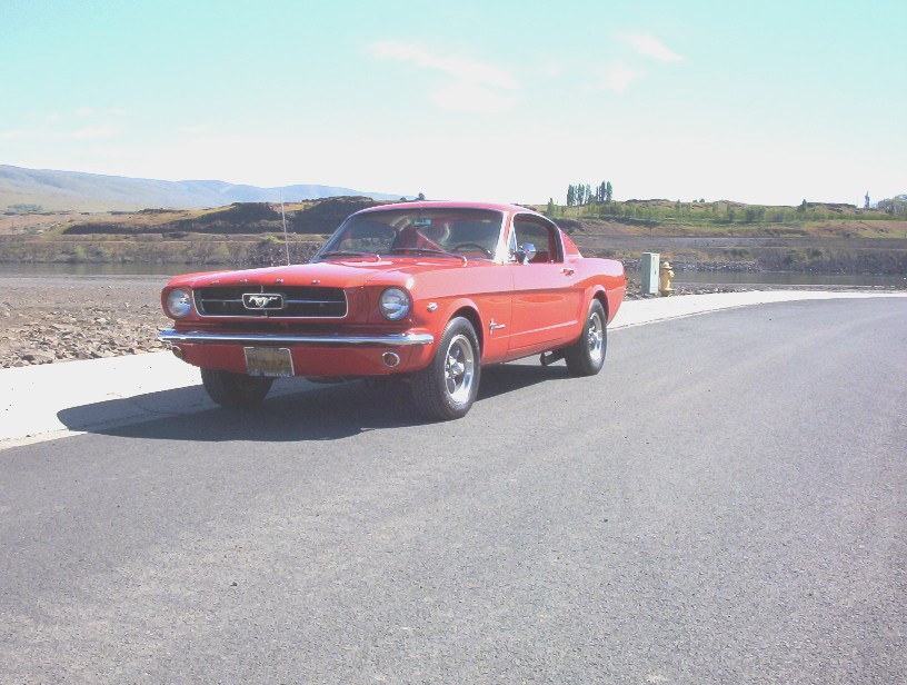 Red classic Ford Mustang fastback, front three-quarter view on a road near water and hills