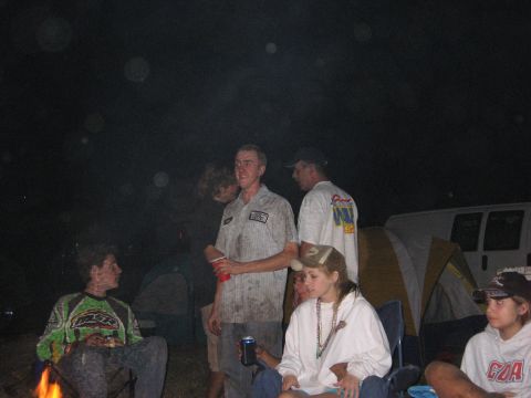 1974 Ford F-100 - A group of people are gathered around a campfire at night, with tents and a van visible in the background.