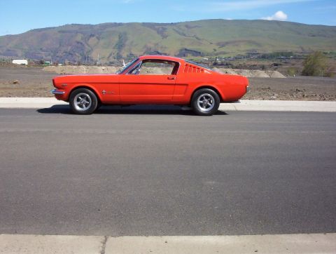 1965 Ford Mustang - A bright red 1965 Ford Mustang coupe is parked on the side of a road with a hilly landscape behind it.