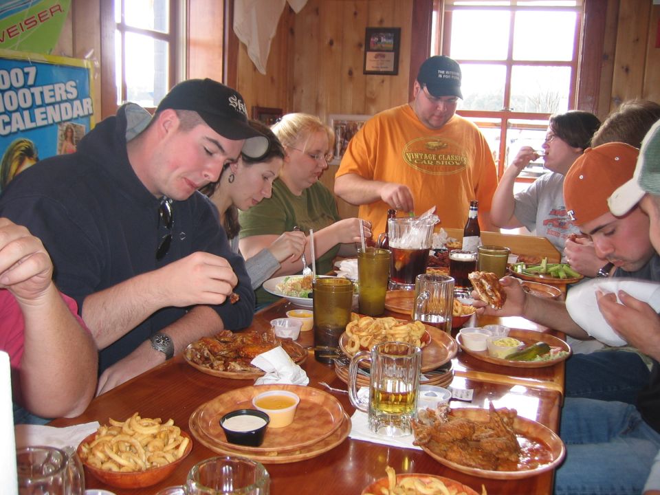 People eating fried chicken, fries, and sandwiches at a wooden table. A 2007 Hooters calendar is visible on the wall